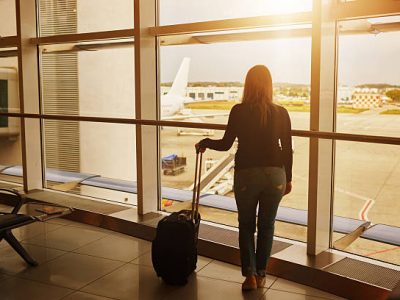 rear view of young woman inside airport looking on the window, sitting near her suitcase and waiting.